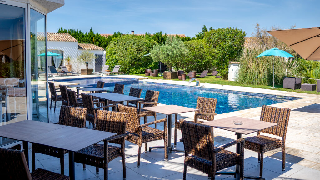 Terrasse de l’hôtel avec tables et chaises, vue sur la piscine avec fontaine, olivier et espace cosy avec bains de soleil, fauteuils et parasols